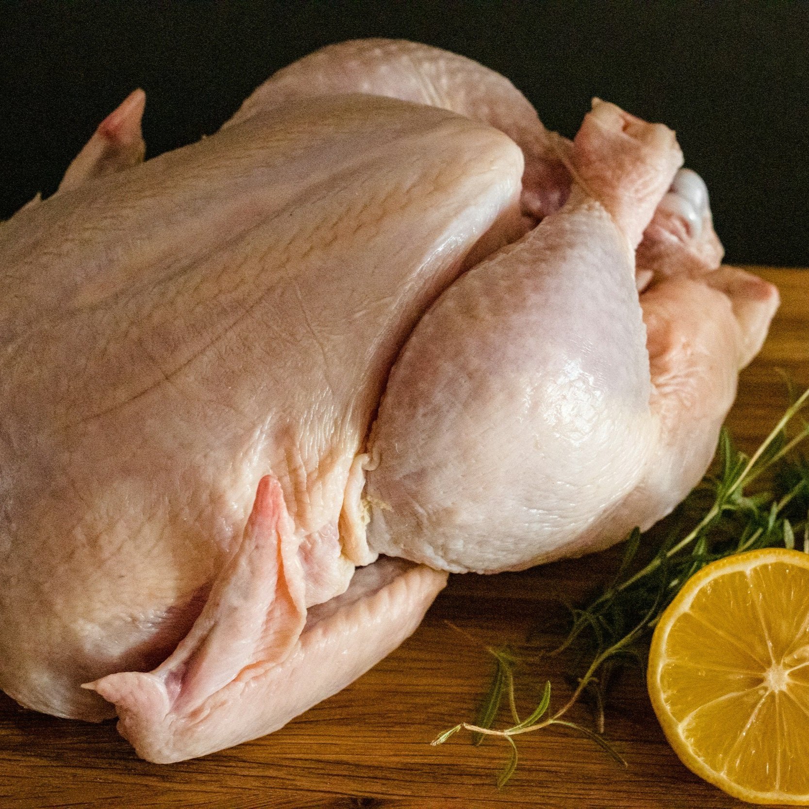 image-asset (2) Raw whole chicken on a wooden cutting board with a slice of lemon and fresh herbs, ideal for cooking or meal prep.